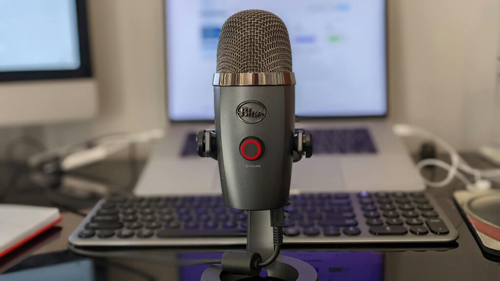 Close-up of a microphone with a blurred keyboard and laptop in a home office setup. Ideal for podcasting content.